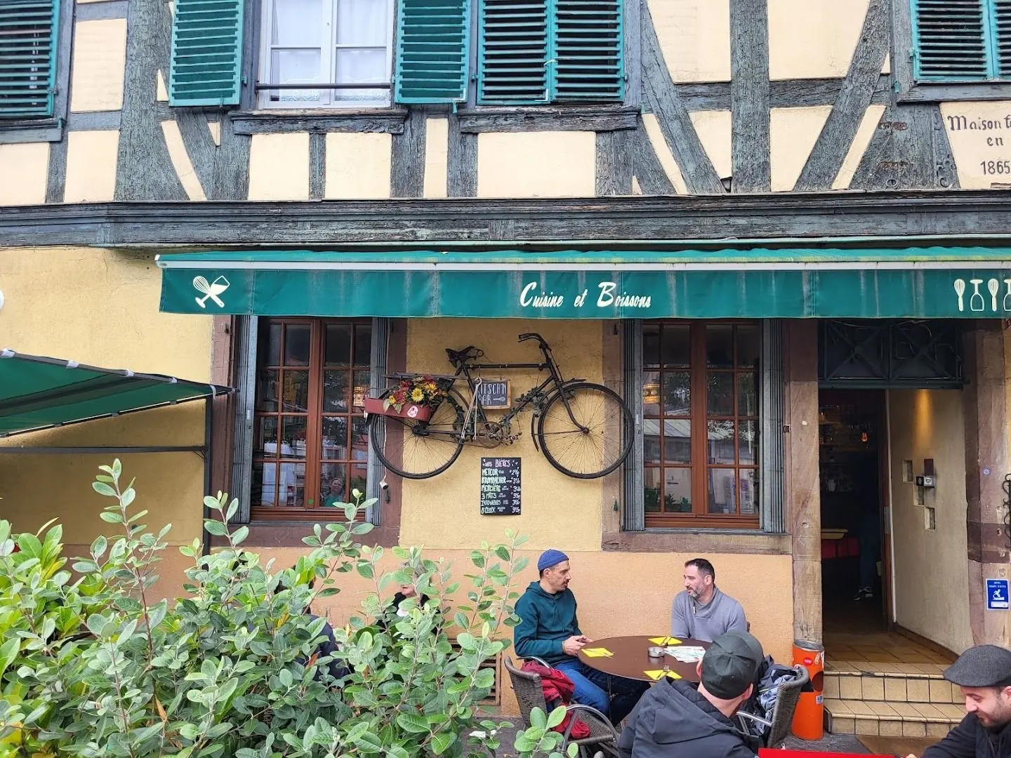 La façade jaune à colombages du Kitsch'n Bar, avec son store vert « Cuisine et Boissons »